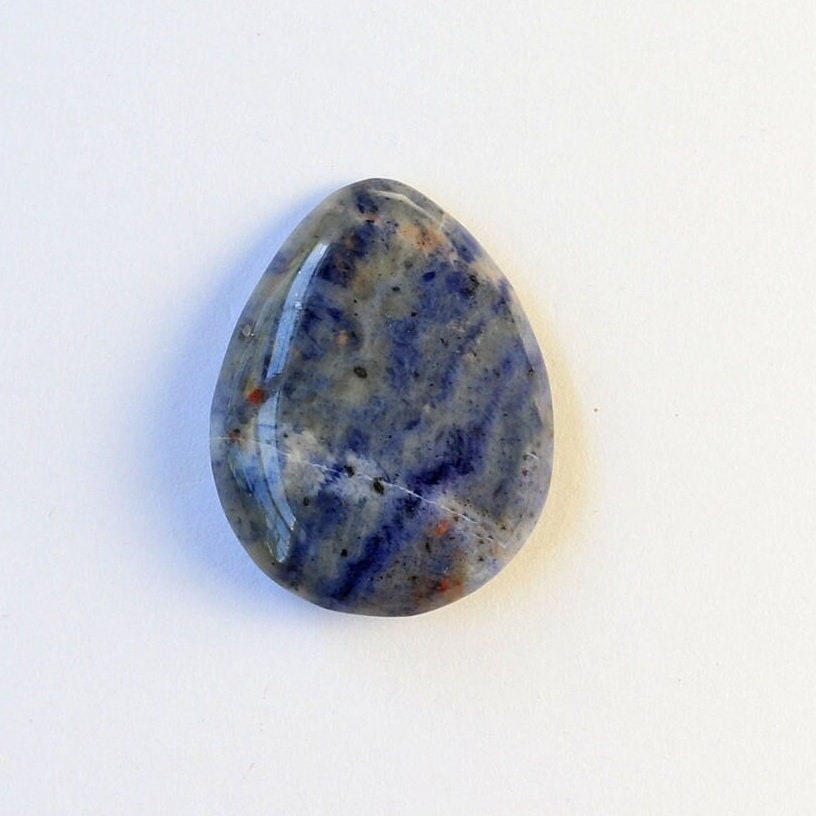 Closeup overhead view of a Blue Foidolite teardrop cabochon, hand-cut and polished in our Missouri studio to showcase its natural blue, gray, cream, and subtle rust-colored patterning.  Shown against white background.
