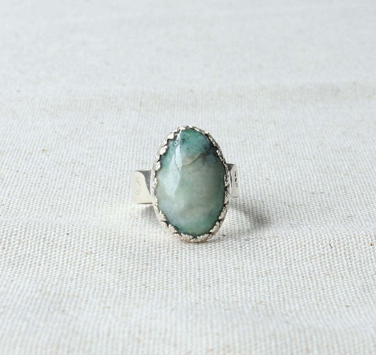 A silver ring with a teardrop-shaped light green stone, set against a textured off-white fabric background.