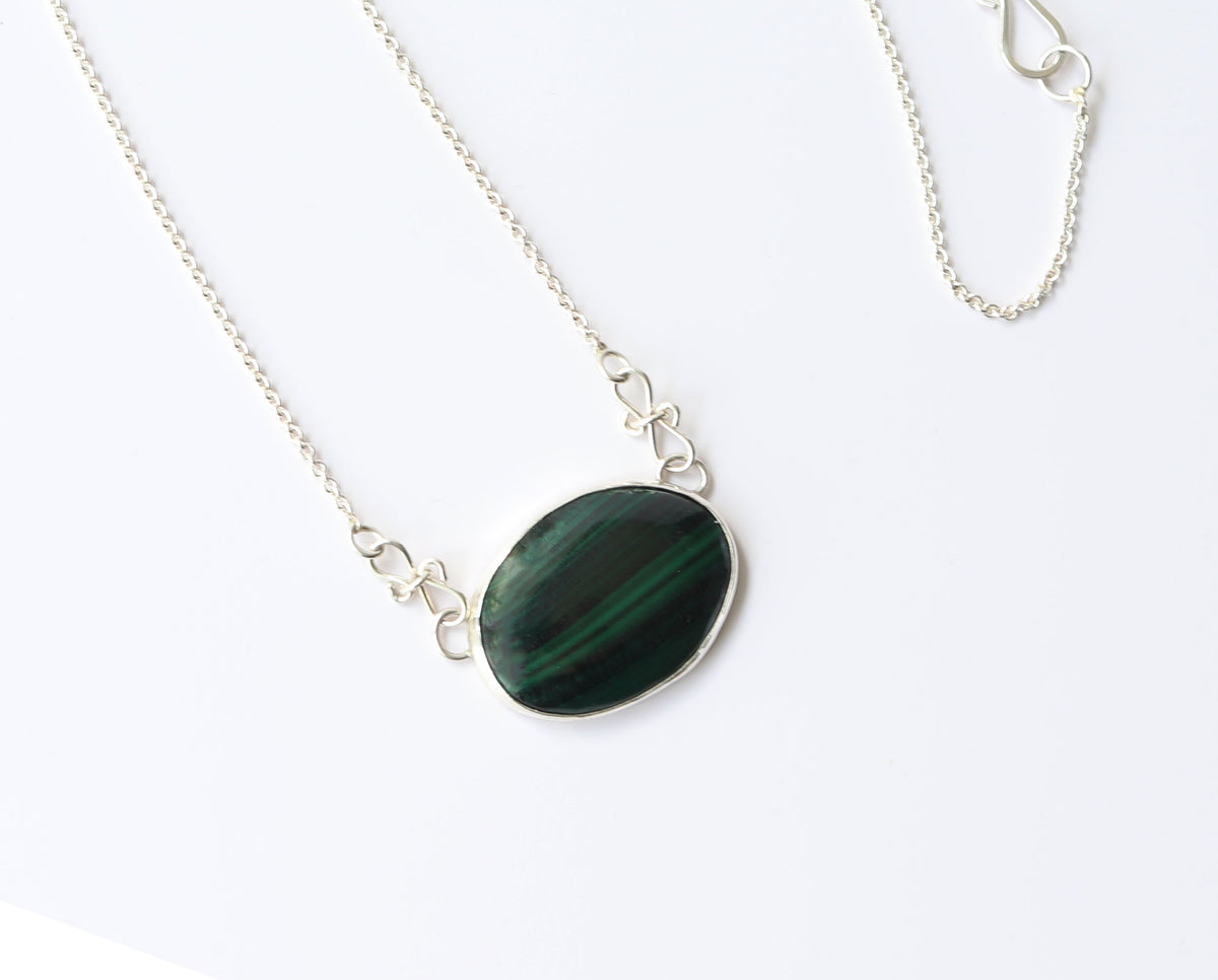 A Malachite gemstone pendant hand-cut and polished stone and set in solid sterling 935 Argentium® Silver. Designed with a custom chain and S- hook for easy wearability. Shown against a white marble background.