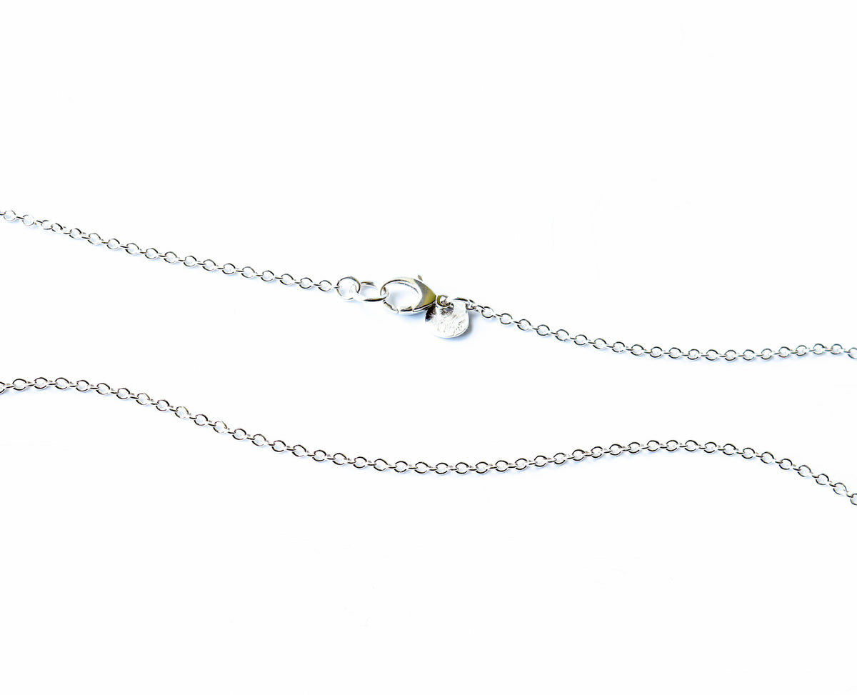A close-up of a delicate silver chain necklace with a lobster clasp and a small round hallmark tag, shown on a plain white background.