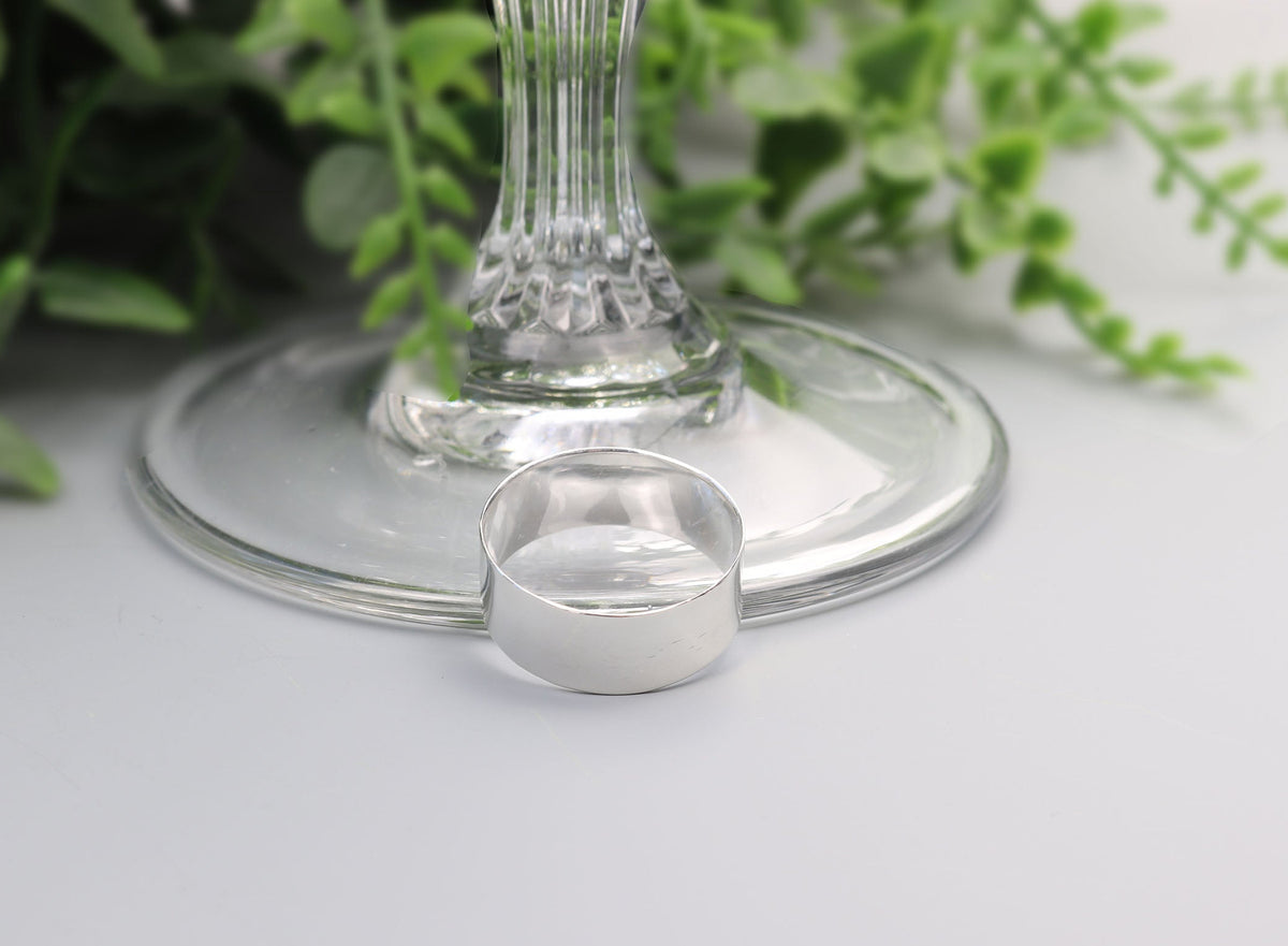A small clear glass teardrop-shaped candle holder or candle cup placed in front of a larger clear glass base, with green foliage blurred in the background on a light gray surface.