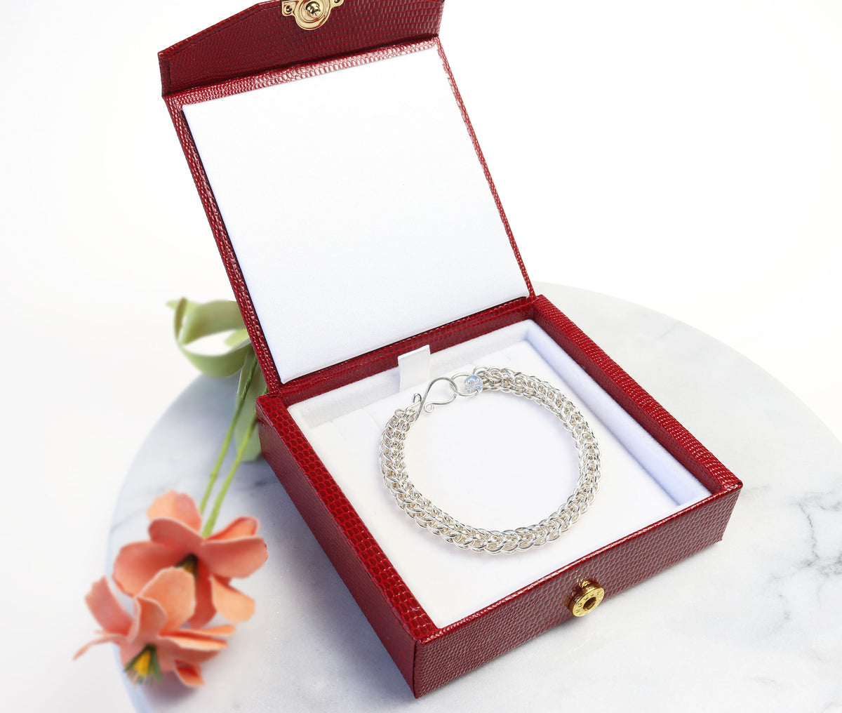 A silver chain bracelet displayed inside an open red textured jewelry box with a white interior, set on a white marble surface with a small peach flower beside it.