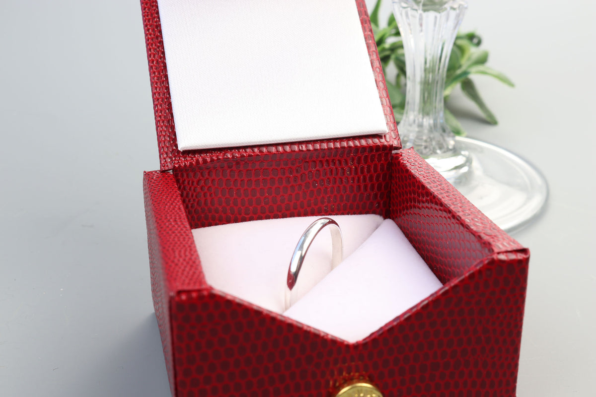 A handcrafted solid Argentium 935 silver half-round ring shown inside an open textured red jewelry box with a white interior, set on a light gray surface with a glass vase in the background.