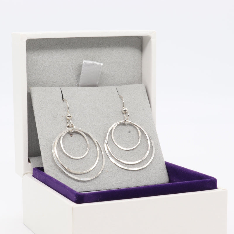 A pair of Argentium silver triple concentric hoop dangle earrings displayed on a gray velvet stand inside an open white jewelry box with a purple velvet base, set against a plain white background.
