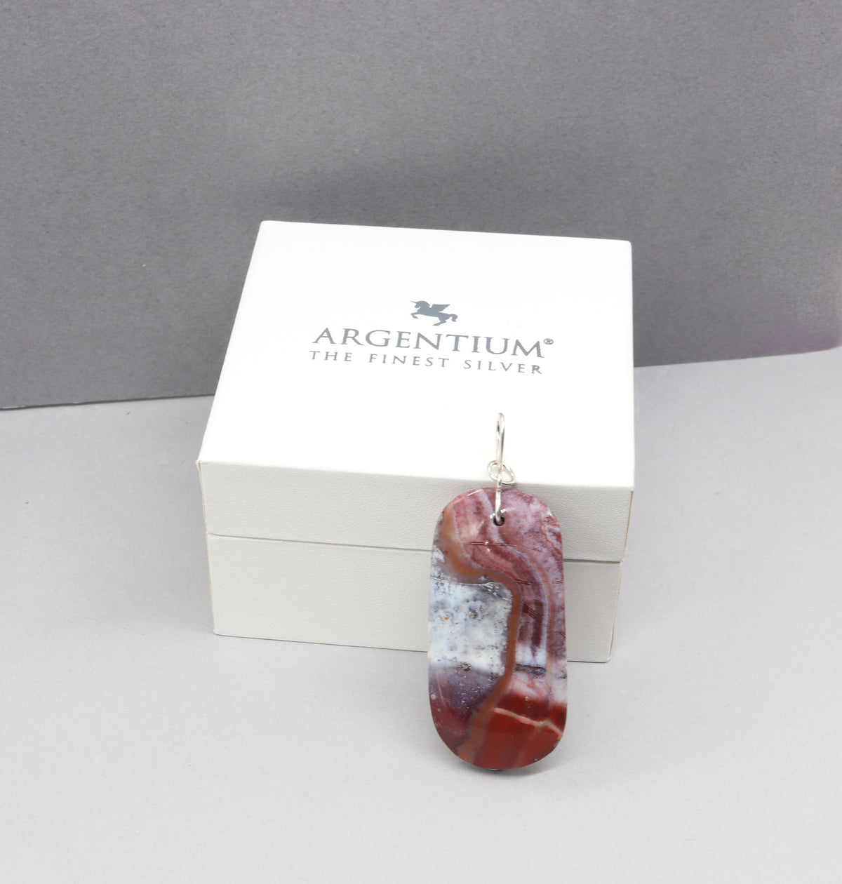An Argentium silver loop with a polished agate pendant featuring red, white, and gray marbled patterns, displayed in front of its white Argentium brand box.