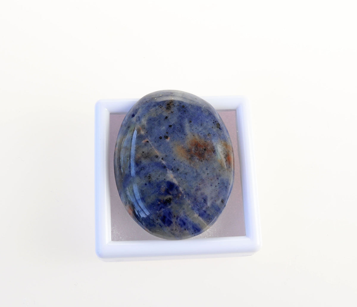 Hand-cut Blue Foidolite Sodalite oval cabochon with mottled blue, creamy white, gray, and warm brown natural patterns.  Shown on a white gem box against a white background.