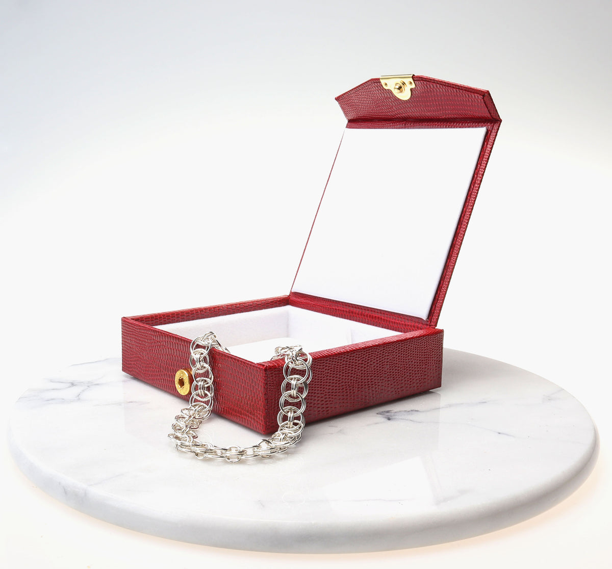 A red textured jewelry box with an open lid, gold-tone clasp, and an Argentium silver helm weave chainmail  bracelet draped over the front, displayed on a white marble round tray against a white studio background.