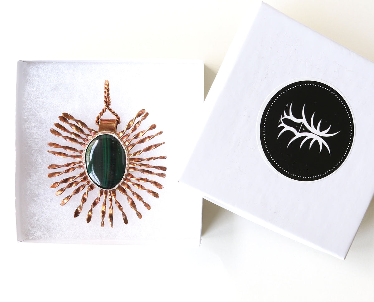 A rose gold sunburst pendant with a dark green malachite oval center, displayed in a white jewelry box next to its lid which features a black circular logo with a white antler design.