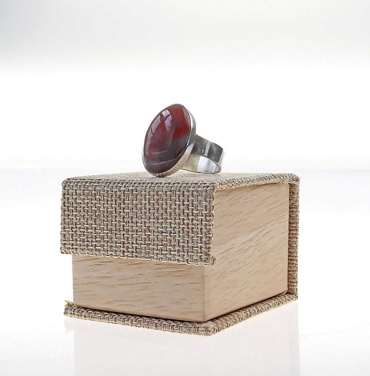 A Handcrafted Petrified Rainbow Wood ring with oval red and dark brown gemstone set in polished Argentium 935 silver, displayed on top of a light wood jewelry box with a woven fabric lid, set against a bright white background.
