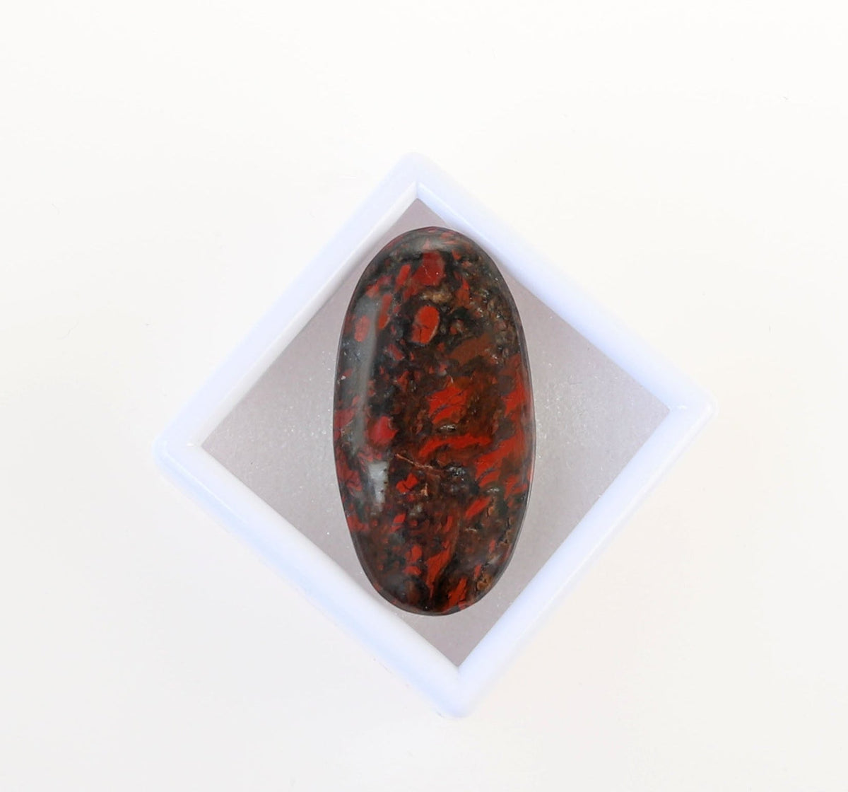 Front view of a handcut oval Red and Black jasper gemstone cabochon, hand-cut and polished into an oval shape to reveal natural mottled patterns of rich reds, earthy blacks and a small white inclusion within the stone.  Shown on a white gem box againt a white background.