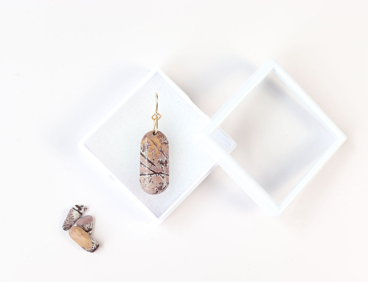 Sonoran Dendritic Rhyolite pendant shown in white gift box on a white background next to a few raw Sonoran Dendtritic pebbles for props.