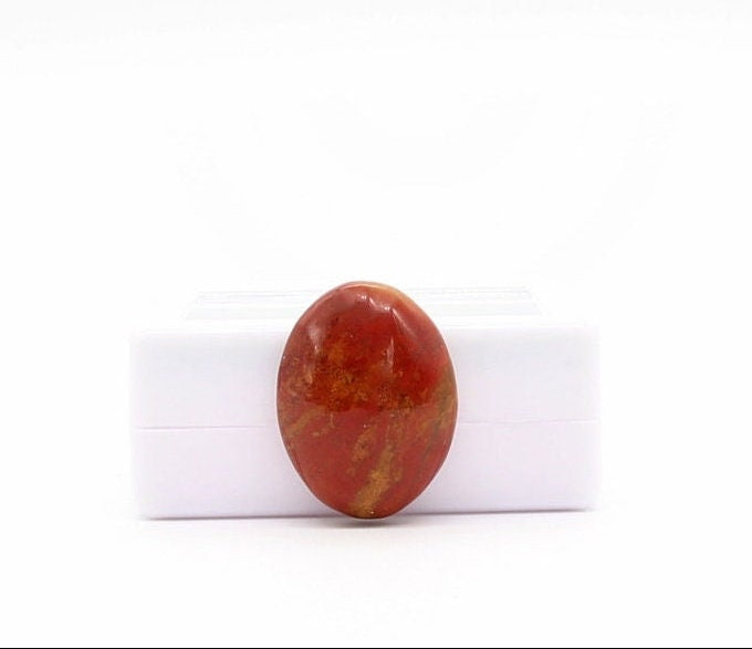 A polished oval Sonoran Sunrise cabochon red stone with golden-yellow marbling, displayed leaning against a small white gem gift box against a white background.