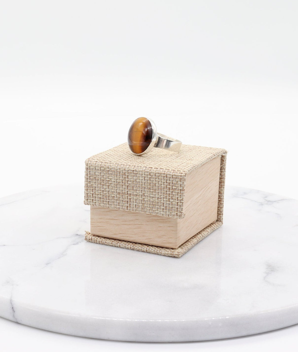 A silver ring with a round brown tiger's eye stone displayed on top of a small woven beige jewelry box, which sits on a round white marble platter against a white background.