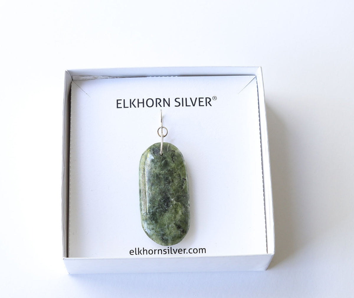A polished green Wyoming Nephrite jad stone pendant with a silver bail, presented inside its white branded gift box by Elkhorn Silver.
