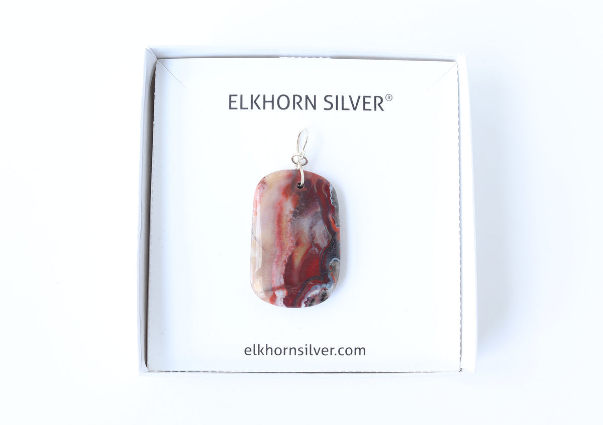 A rectangular Red Mexican pendant with a red, white, and brown agate-like marbled pattern, displayed on a white card inside a white gift box. The card is branded 'ELKHORN SILVER' with the website 'elkhornsilver.