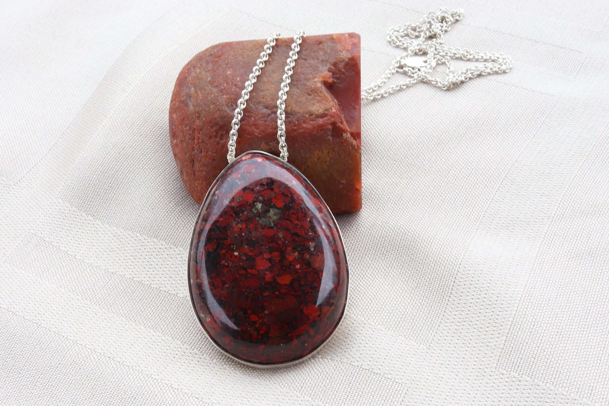 A Brecciated jasper gemstone pendant. Hand-cut and polished into an oval shape to reveal natural hues of rich reds, earthy browns, subtle grays, and delicate hints of creamy white.  The stone is set in solid 935 Argentium Silver and includes a complementary 935 silver chain.