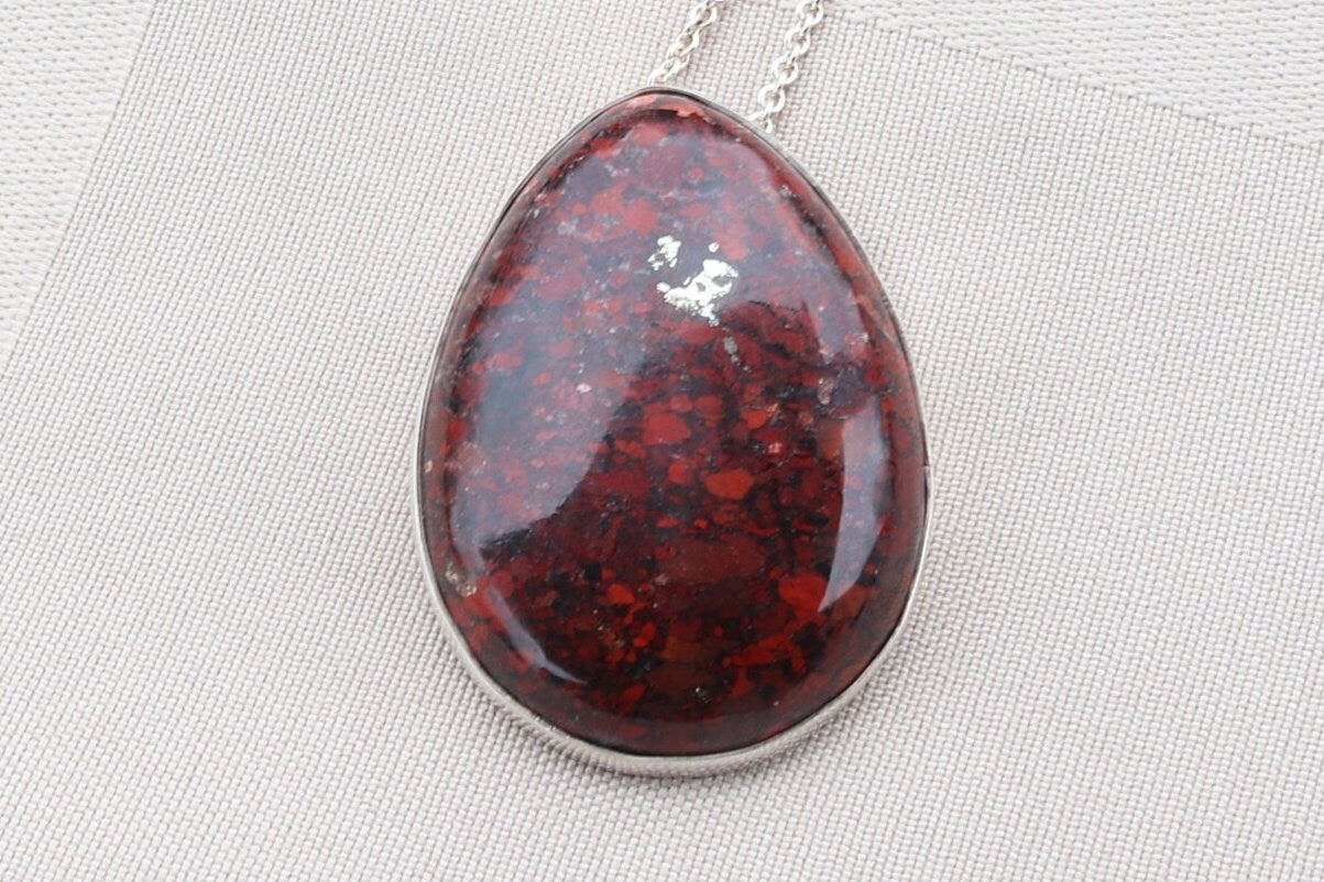 A Brecciated jasper gemstone pendant. Hand-cut and polished into an oval shape to reveal natural hues of rich reds, earthy browns, subtle grays, and delicate hints of creamy white.  The stone is set in solid 935 Argentium Silver and includes a complementary 935 silver chain.