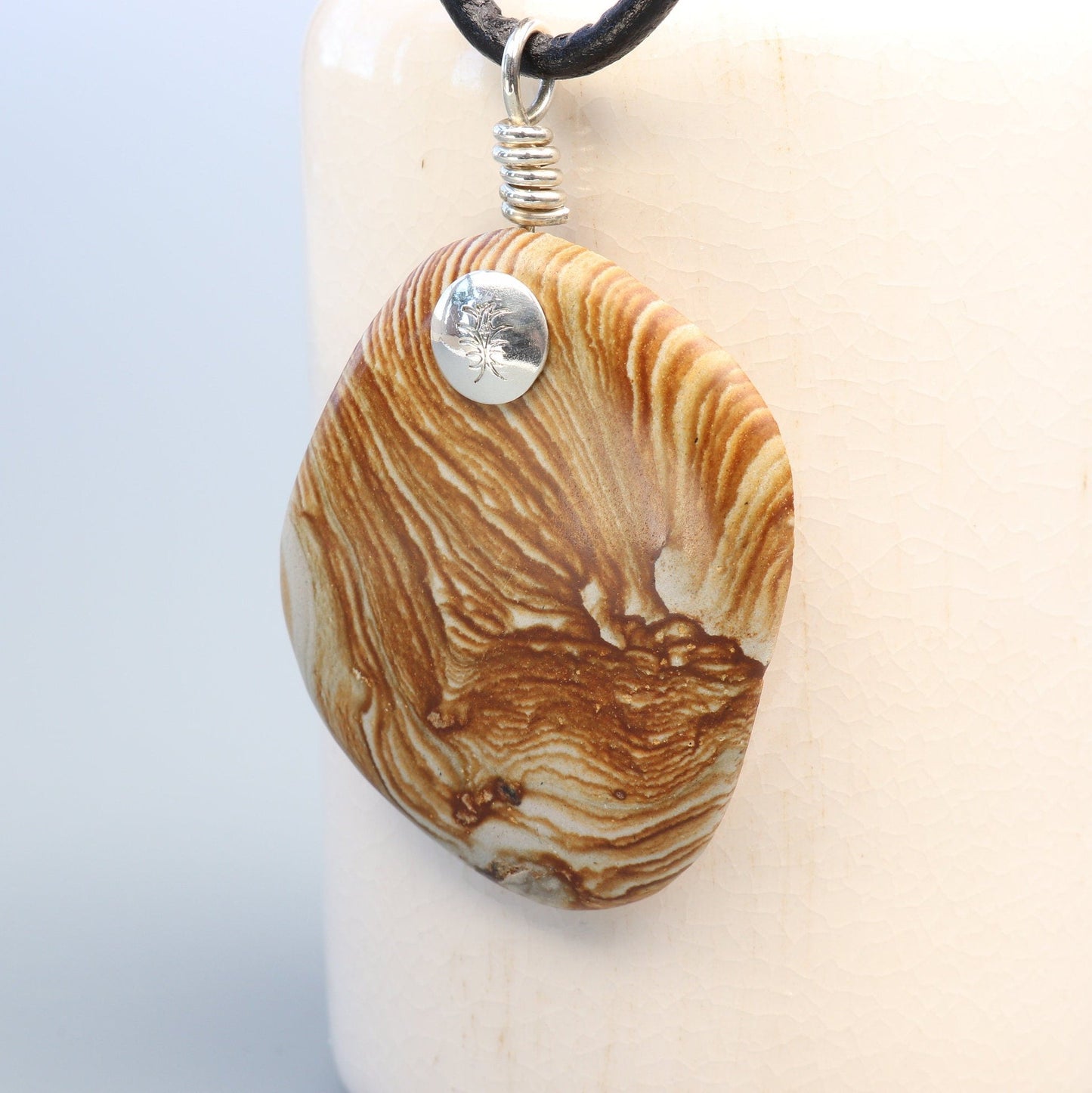 A natural Picture Jasper gemstone, handcut and polished on both sides to showcase the earthy tones, warm browns, creamy beiges, and nature's intricate patterns.  The custom handcrafted coil bail is from sterling Argentium® 935 Silver renowned for its resistance to tarnishing and its durable quality.
To complete the look a complementary 3 mm Leather Cord Necklace is included.