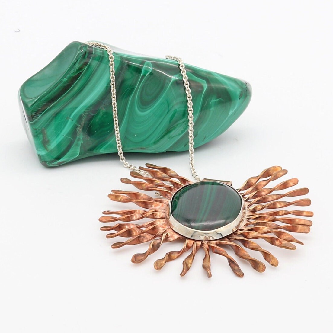 A unique natural Malachite Gemstone Pendant handcut and polished into a cabochon and encased in a sterling 935 Argentium silver setting surrounded with a unique starburst pattern in Jewelers Brass with a captivating copper patina. Plus to complete the look a complementary sterling 935 Argentium silver chain is included.