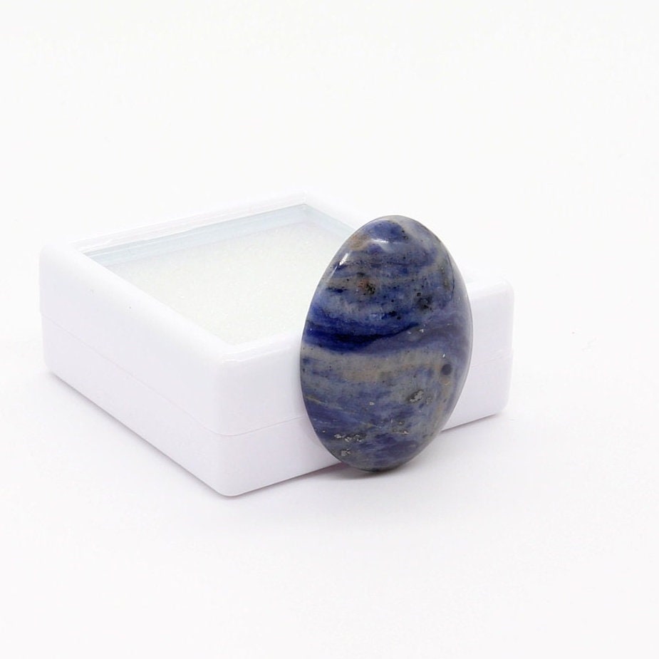 A hand-cut and polished natural Foidolite cabochon with layered deep blue, slate blue and gray natural mineral patterning, displayed at a left angle against a white gem box, photographed on a clean white background.