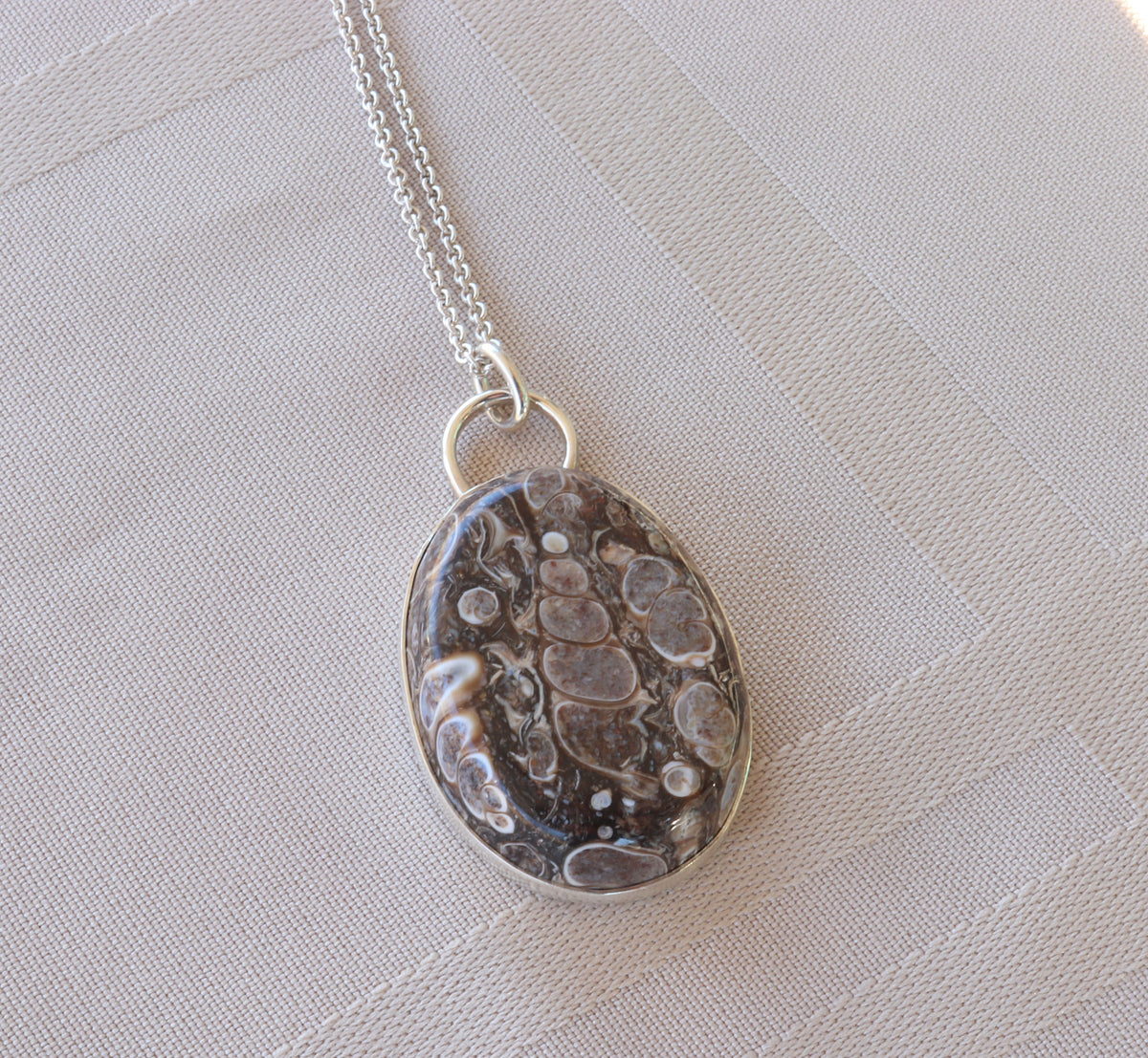 Natural Turritella Agate fossil pendant in Argentium 935 Silver with silver chain, displayed on a textured beige fabric background.