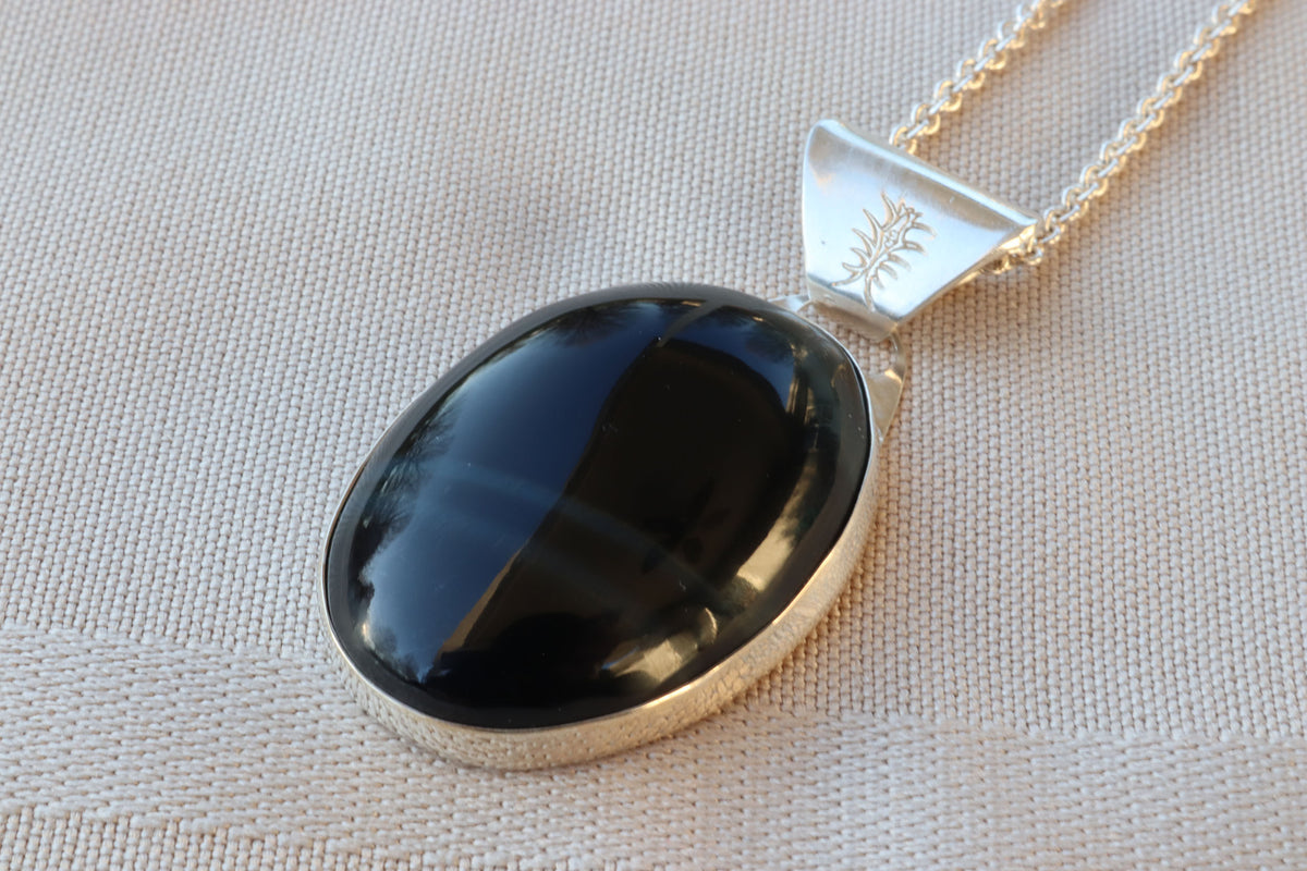 An oval natural Blue Tiger’s Eye pendant in a polished Argentium 935 silver bezel, suspended from a silver chain against a white background, resting on a textured beige fabric background.