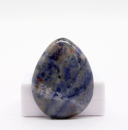 A Blue Foidolite teardrop cabochon, hand-cut and polished in our Missouri studio to showcase its natural blue, gray, cream, and subtle rust-colored patterning.  Shown against a white gem box against white background.