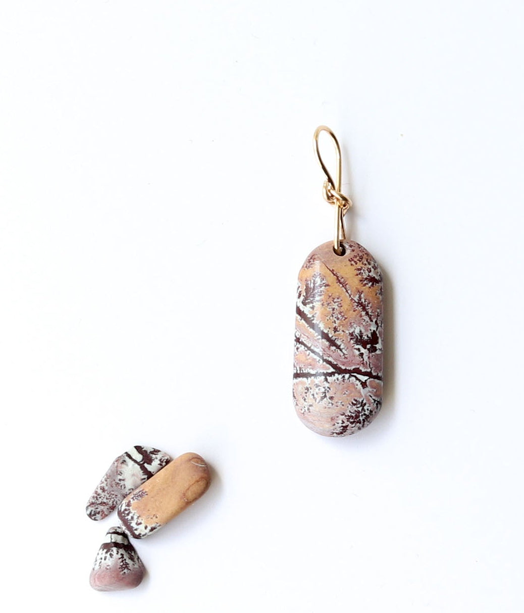 A one-of-a-kind Sonoran Dendritic Rhyolite pendant charm showcases warm tan and soft pink tones laced with delicate white and deep brown dendritic patterns finished with a custom 14/20 gold-filled infinity bow bail.  Shown on white background next a few raw Sonoran Dendritic pebbles for props.