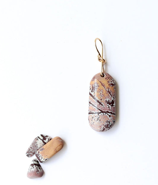 A one-of-a-kind Sonoran Dendritic Rhyolite pendant charm showcases warm tan and soft pink tones laced with delicate white and deep brown dendritic patterns finished with a custom 14/20 gold-filled infinity bow bail.  Shown on white background next a few raw Sonoran Dendritic pebbles for props.