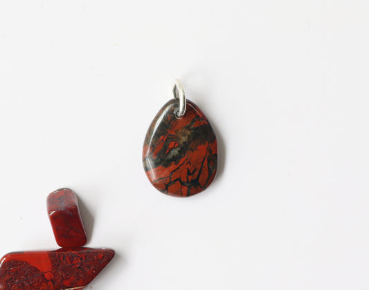 A Red Brecciated Jasper Pendant – featuring a natural mosaic of deep reds, earthy browns, and bold black veining.  Polished on both sides for a reversible finish, this teardrop gemstone offers a smooth feel and versatile wear. Subtle quartz or chalcedony inclusions may be visible, adding natural dimension and authenticity.  Shown on white background.