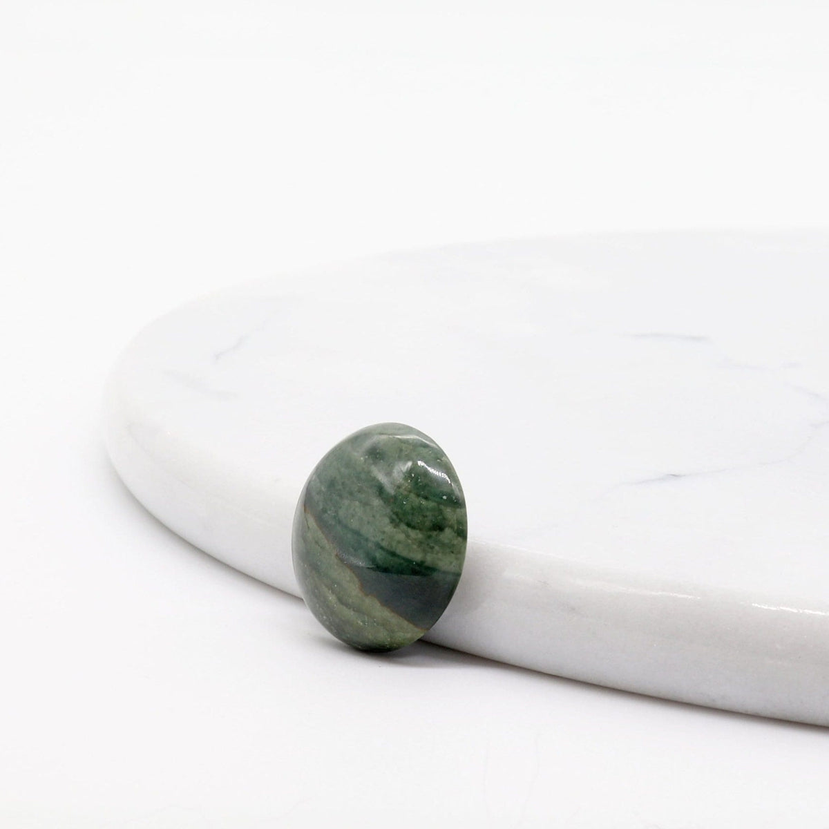 A sideview of this handcut and Polished this Arizona Green Lizard gemstone cabochon features a rich blend of deep green tones and earthy patterns. The stone is shaped and polished into a smooth cabochon shown leaning against white marble slab on a white background.