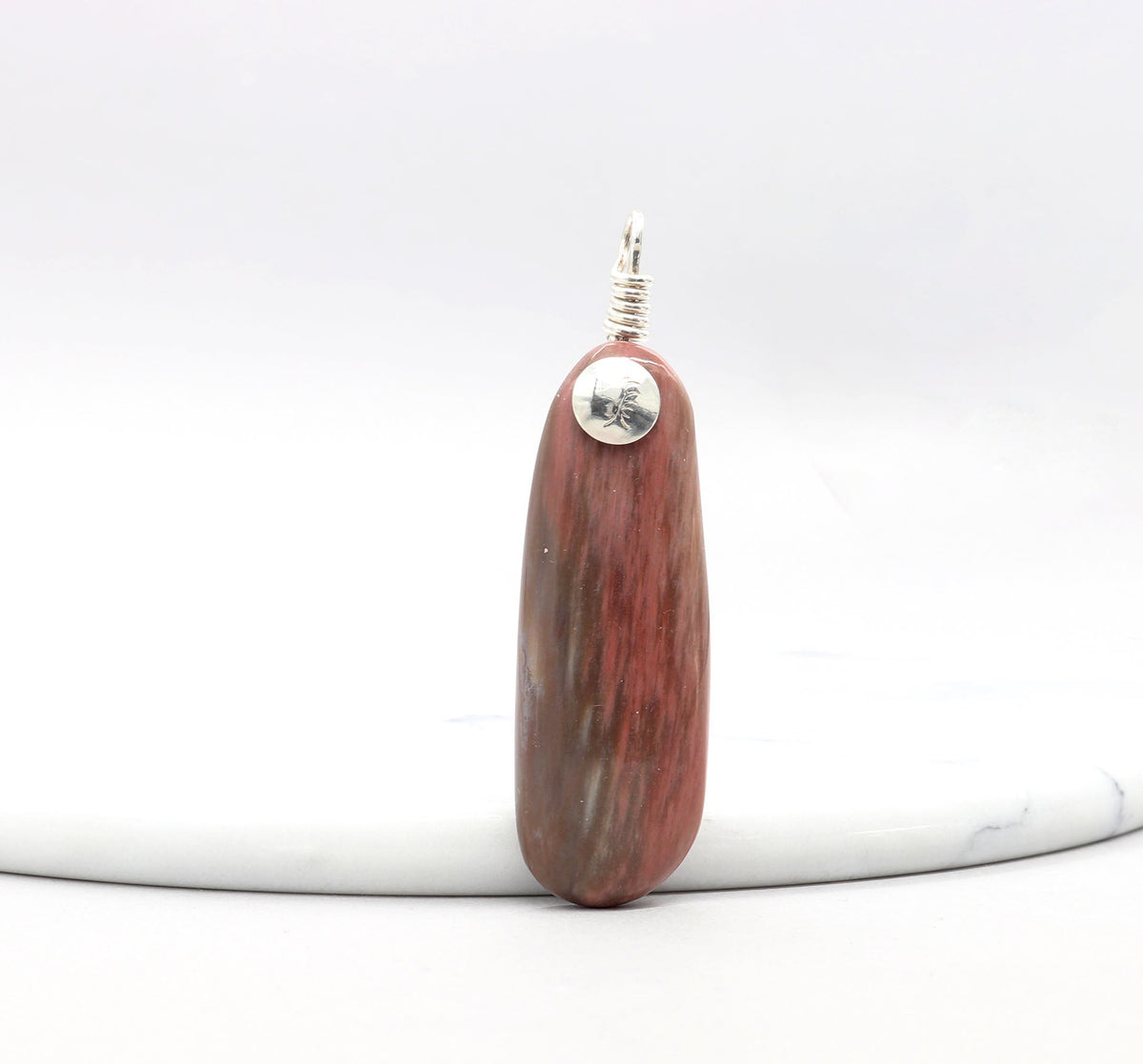 A polished Petrified Wood reddish-brown wooden pendant with Argentium silver accents and wire loop, standing upright on a white marble round platform against a white background.