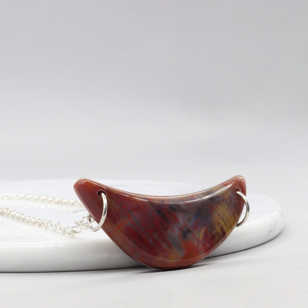 Argentium Necklace with a Petrified Rainbow wood crescent pendant resting at a left angle against a white marble slab on a white background