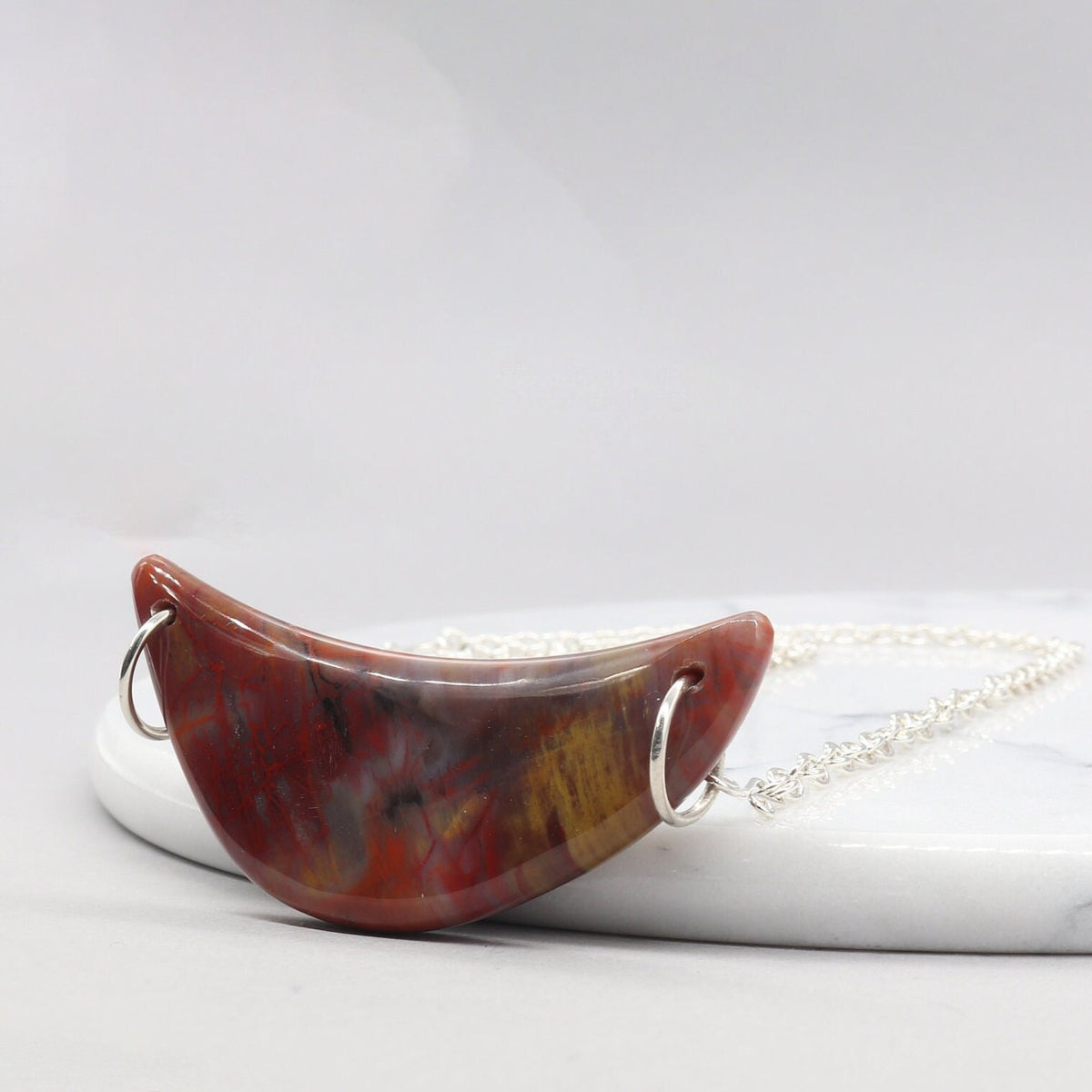 Argentium Necklace with a Petrified Rainbow wood crescent pendant resting at an angle against a white marble slab on a white background