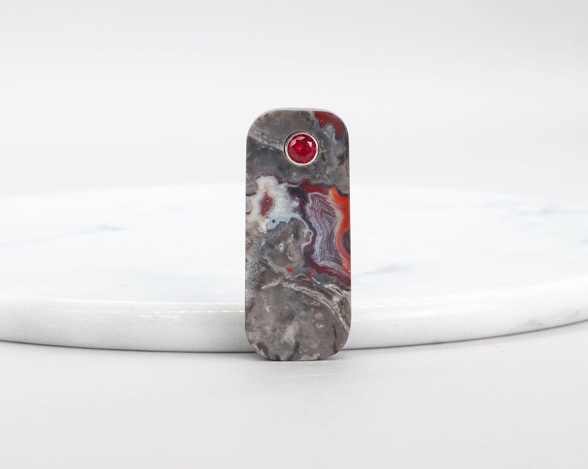 Red Mexican Agate pendant with 6mm lab-grown ruby inlay and Argentium Silver bail shown against a white marble slab on white background.