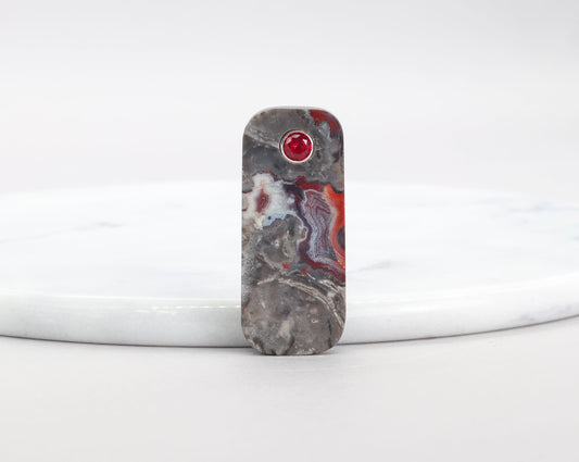 Red Mexican Agate pendant with 6mm lab-grown ruby inlay and Argentium Silver bail shown against a white marble slab on white background.