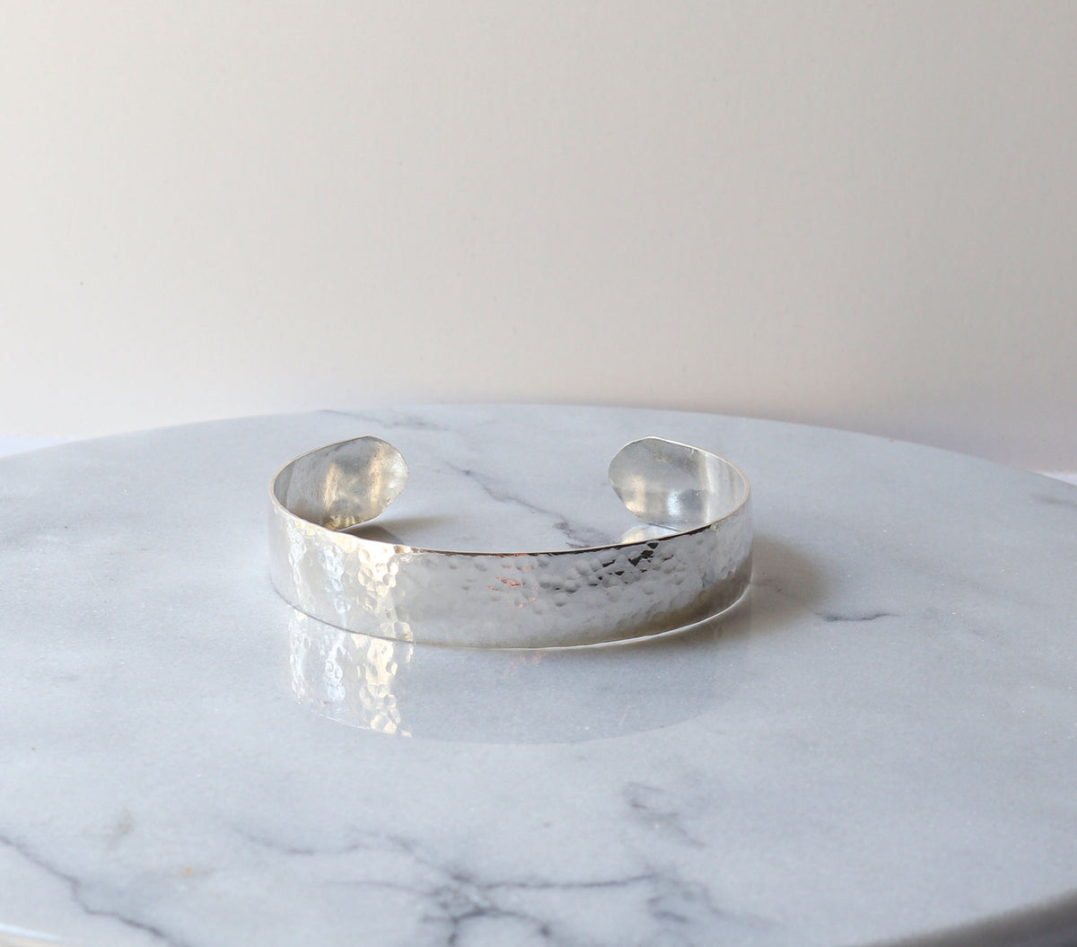Hand-hammered sterling silver cuff bracelet with a softly curved profile, resting on a light marble-patterned surface against a light beige background.