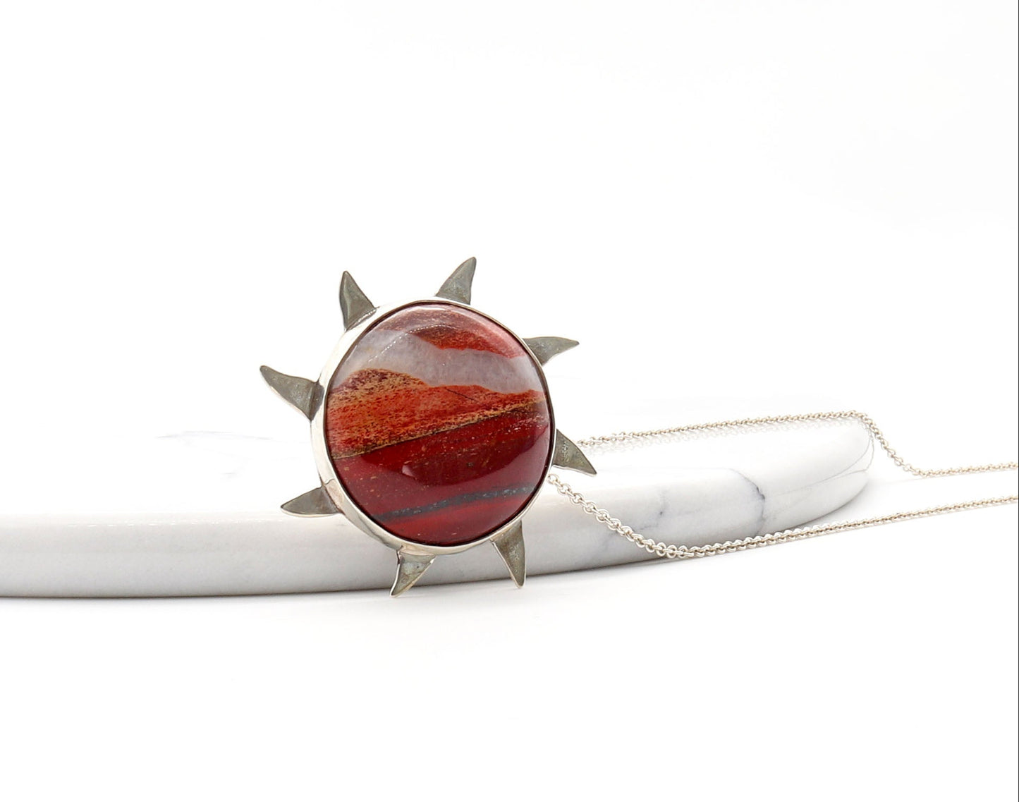 Snakeskin Jasper pendant, hand-cut and polished to showcase its vibrant flaming red, rich brown, and creamy natural patterns. Encased in premium Argentium® 935 silver shown against white marble slab with Argentium necklace