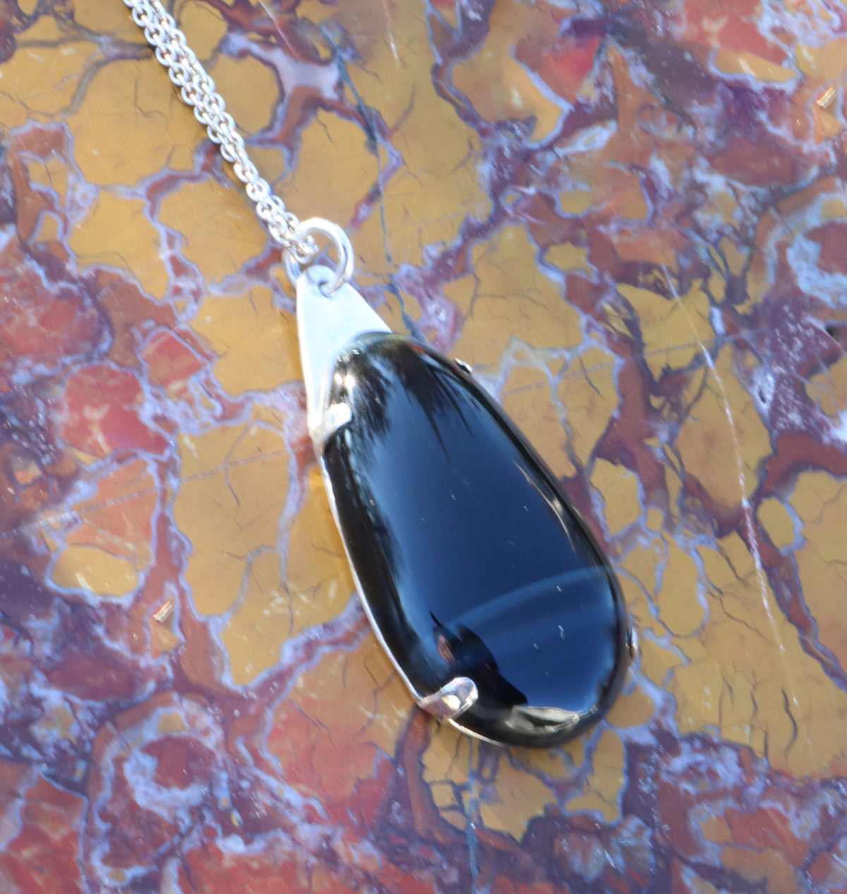 A Handcrafted African Blue Tiger's Eye teardrop pendant with deep blue-black reflective gemstone, solid Argentium 935 silver tabbed setting and silver chain., resting on a textured, marbled surface with yellow, red, and brown tones.