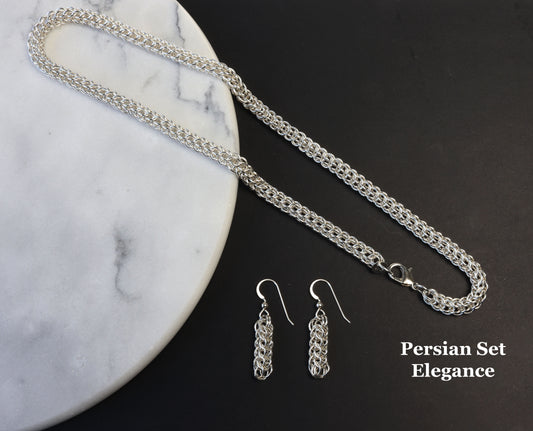 A stunning Full Persian Weave necklace, handmade from over 400 precision-cut rings of premium 935 Argentium® Silver, this necklace offers exceptional tarnish resistance and hypoallergenic properties, making it a superior choice for both durability and everyday elegance. Shown on white marble with contrasting black background with matching earrings.