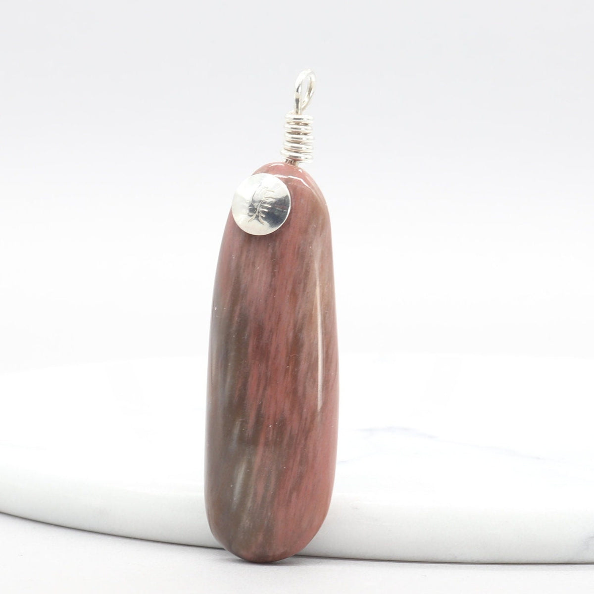  A polished Petrified Wood reddish-brown wooden pendant with Argentium silver accents and wire loop with makers mark stamped on front of metal, standing upright at right angle on a white marble round platform against a white background.