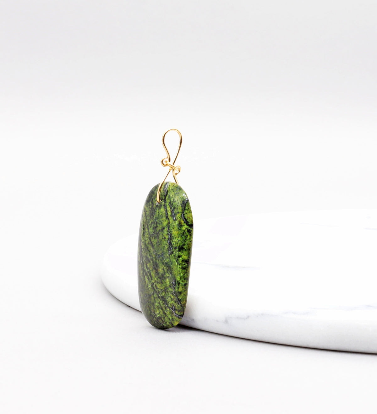 A Forest Green Jasper pendant with a gold filled wire loop, resting on a white marble slab against a white background.