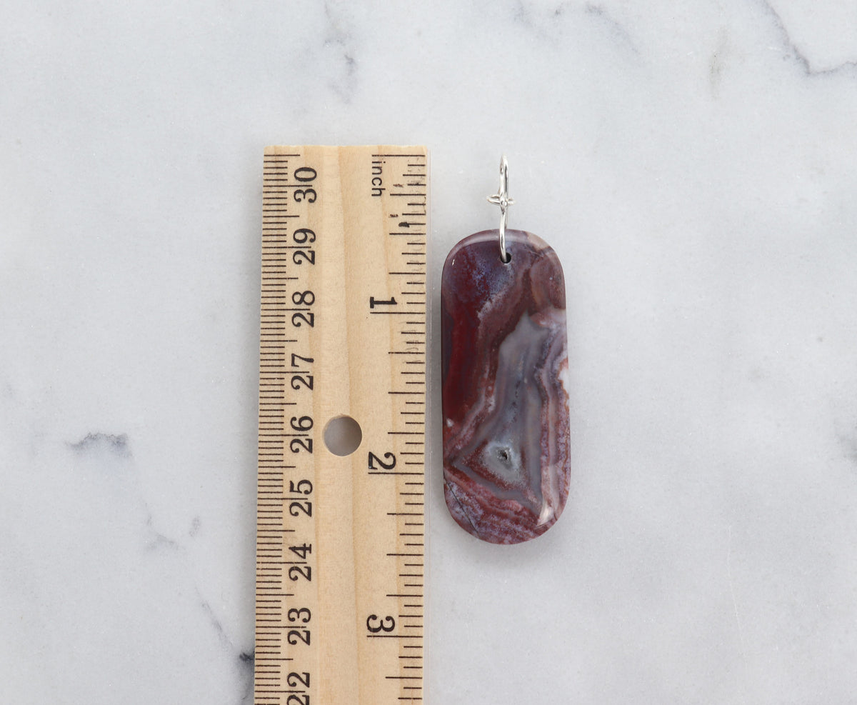 A polished Red Mexican agate pendant with a natural banded pattern in shades of mauve-gray and lavender hues, shown next to a wooden ruler on a white marble surface to indicate its size.