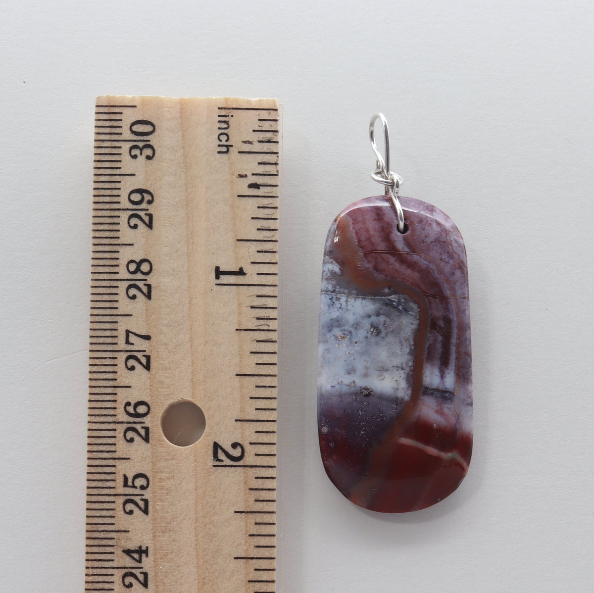 A polished agate pendant with a natural marbled pattern of red, brown, and white, shown next to a wooden ruler for scale on a light gray background.