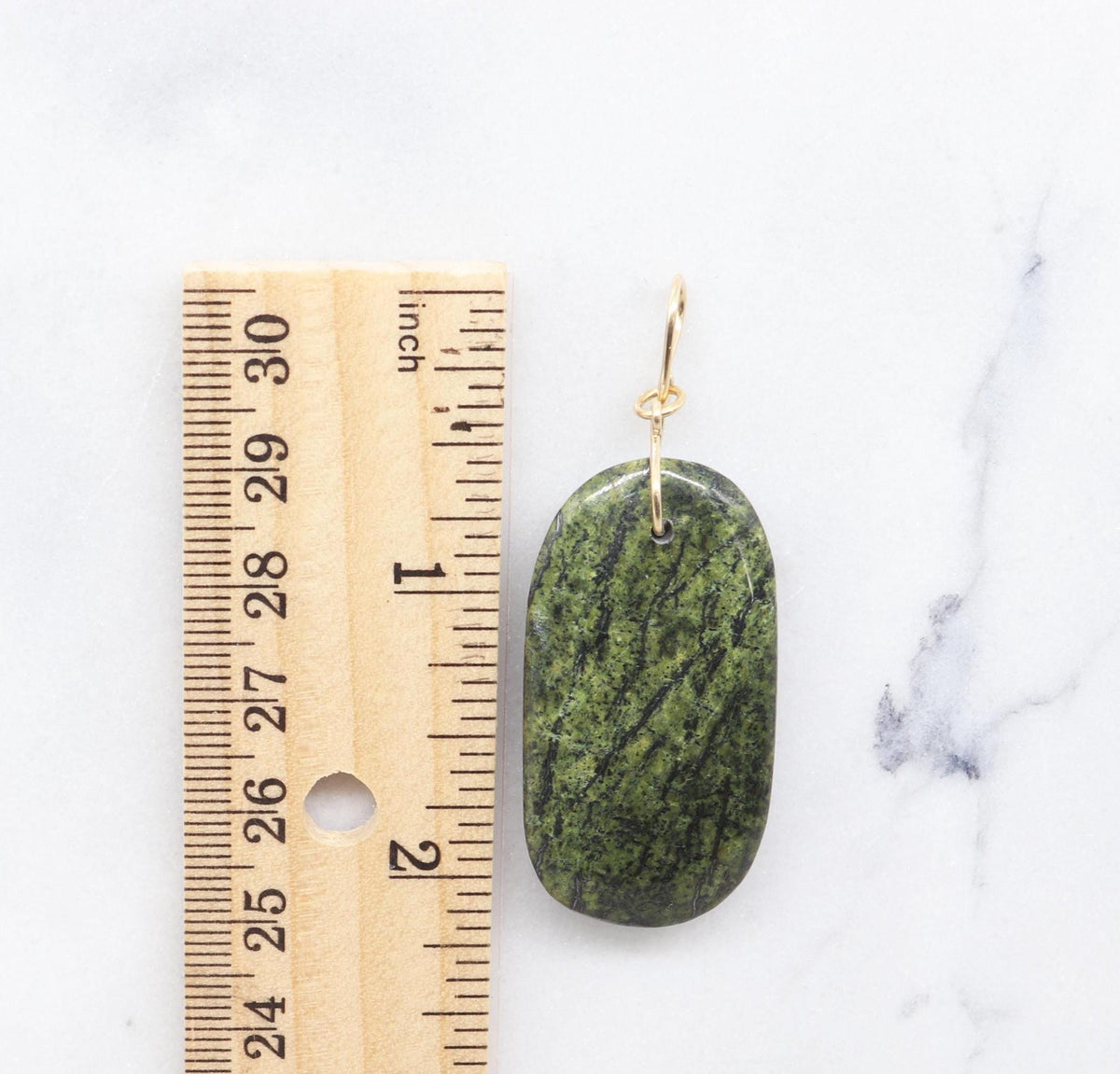 A Green forest Jasper handcut and polished pendant shown next to a wooden ruler for scale on a white marble slab background.