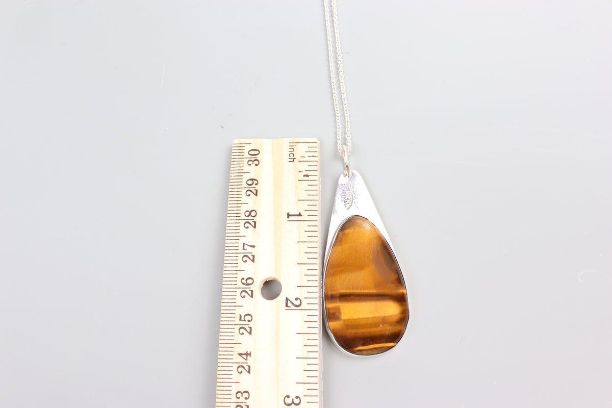 A handcrafted Tiger's Eye Pendant polished to bring out its unique silky wavy brilliance and natural chatoyancy. Set in a custom Argentium® .935 Silver bezel and paired with a complementary silver 935 Argentium necklace chain shown next to ruler for size reference on a light gray background.