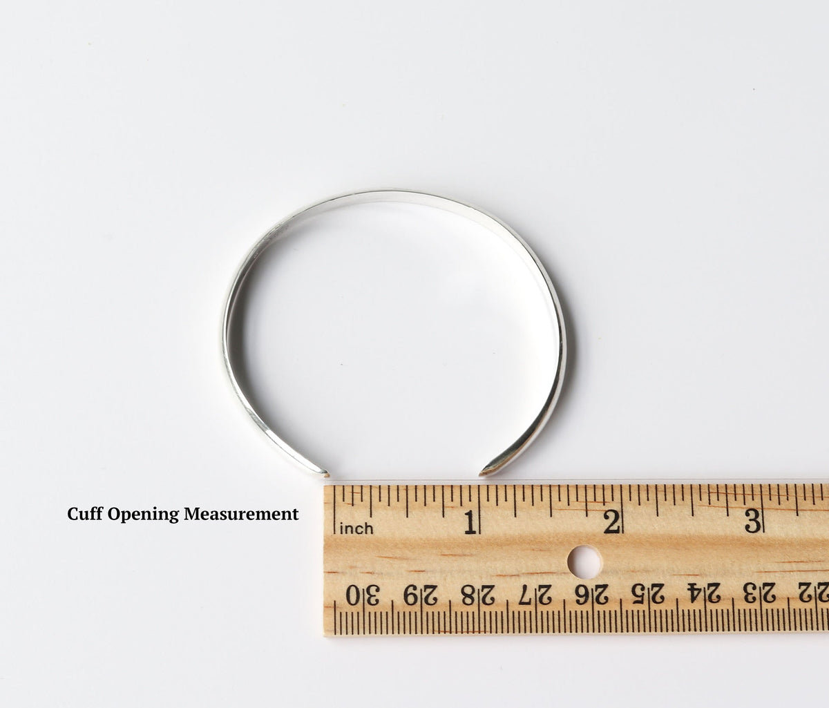 Silver cuff bracelet with wooden ruler demonstrating how the cuff opening is measured.  Shown on a light gray background.
