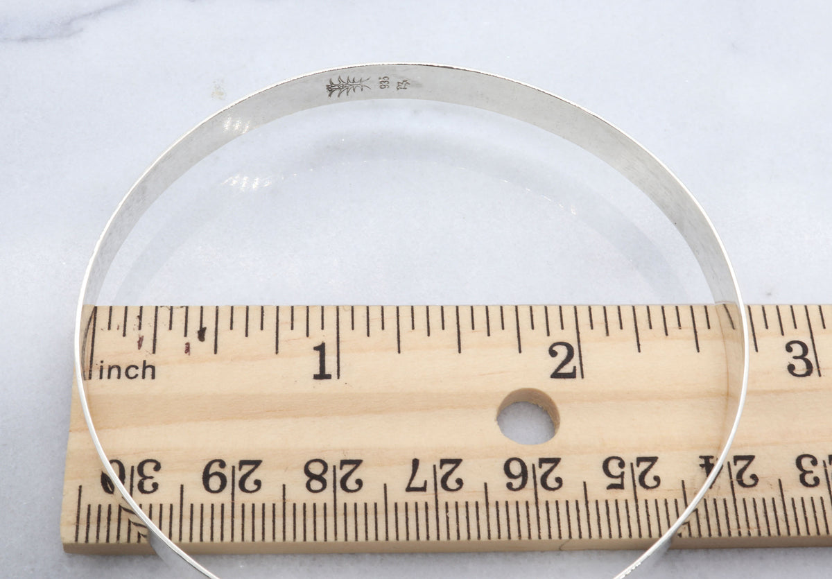 A high Flat Bangle Bracelet in Argentium® 935 silver.   Inside Diameter measures 2.56 inches next to a wooden ruler with inch markings to indicate size, placed on a light marble-like surface.