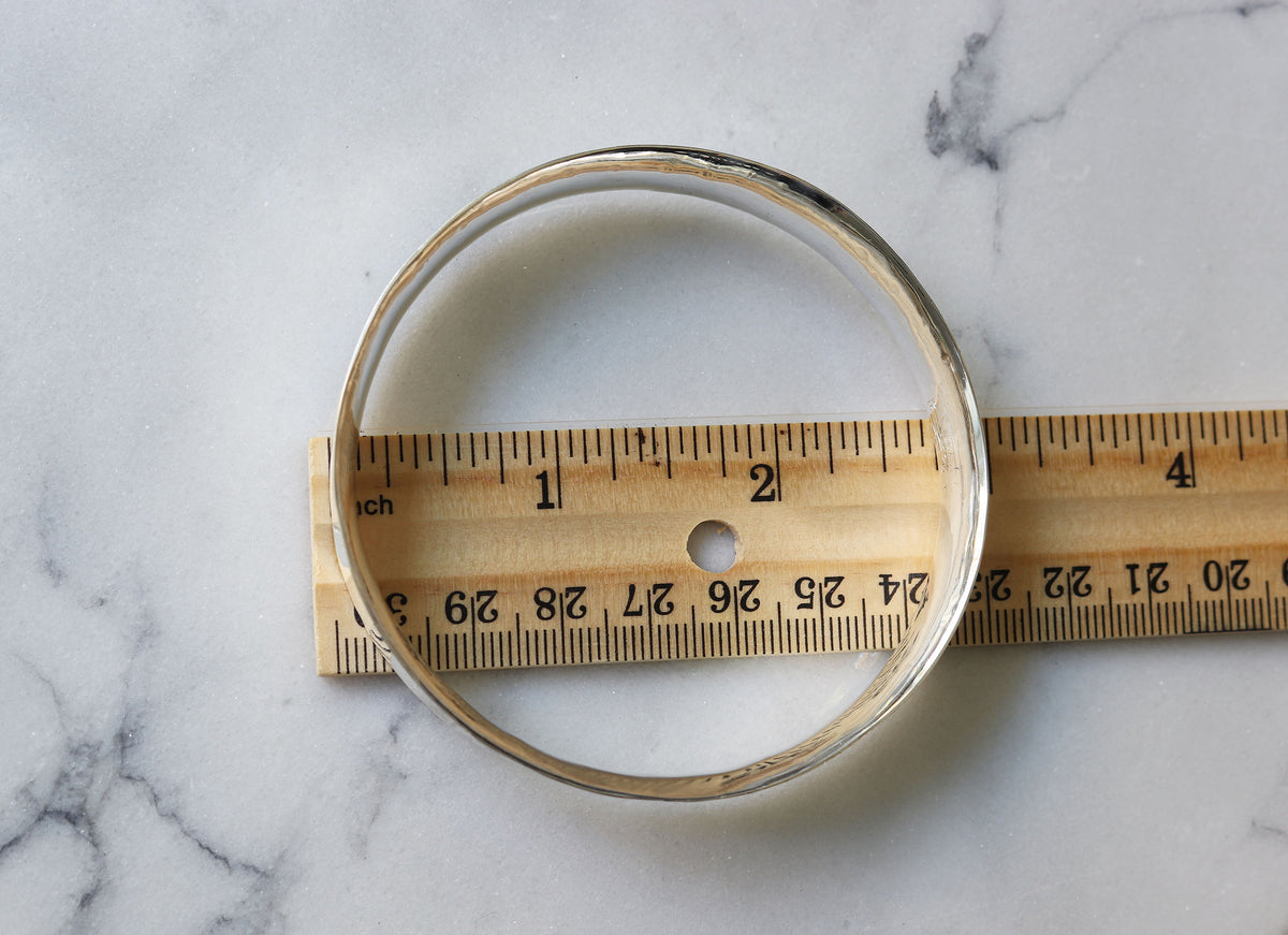 A silver-toned metal bangle bracelet is shown alongside a wooden ruler on a white marble surface to indicate its size.