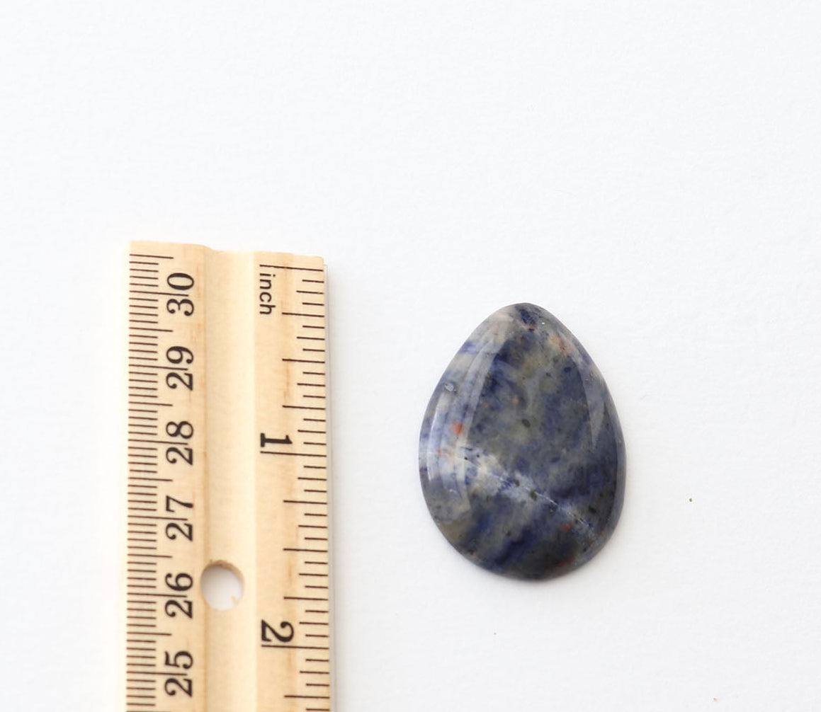 A Blue Foidolite cabochon polished teardrop-shaped with dark blue, gray, and white marbling, shown next to a wooden ruler on a plain white background.